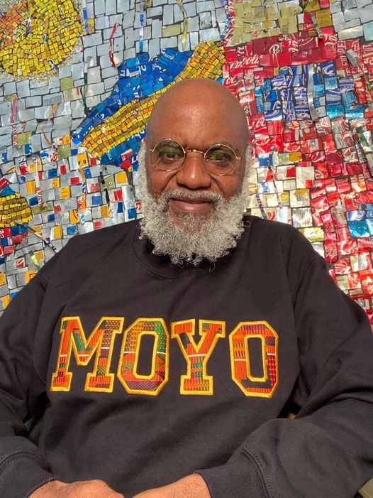 a picture showing moyo okediji wearing a customized hoodie poised for the camera and behind him is one of his art piece
